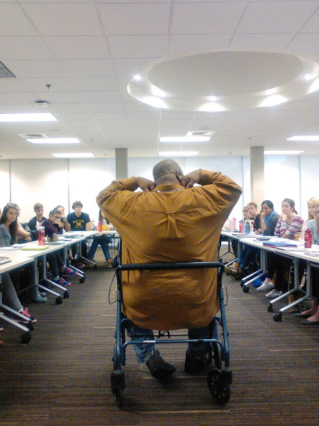 Homeless advocate Steve Thomas sitting in a walker flexing his muscles as he speaks to a group of students